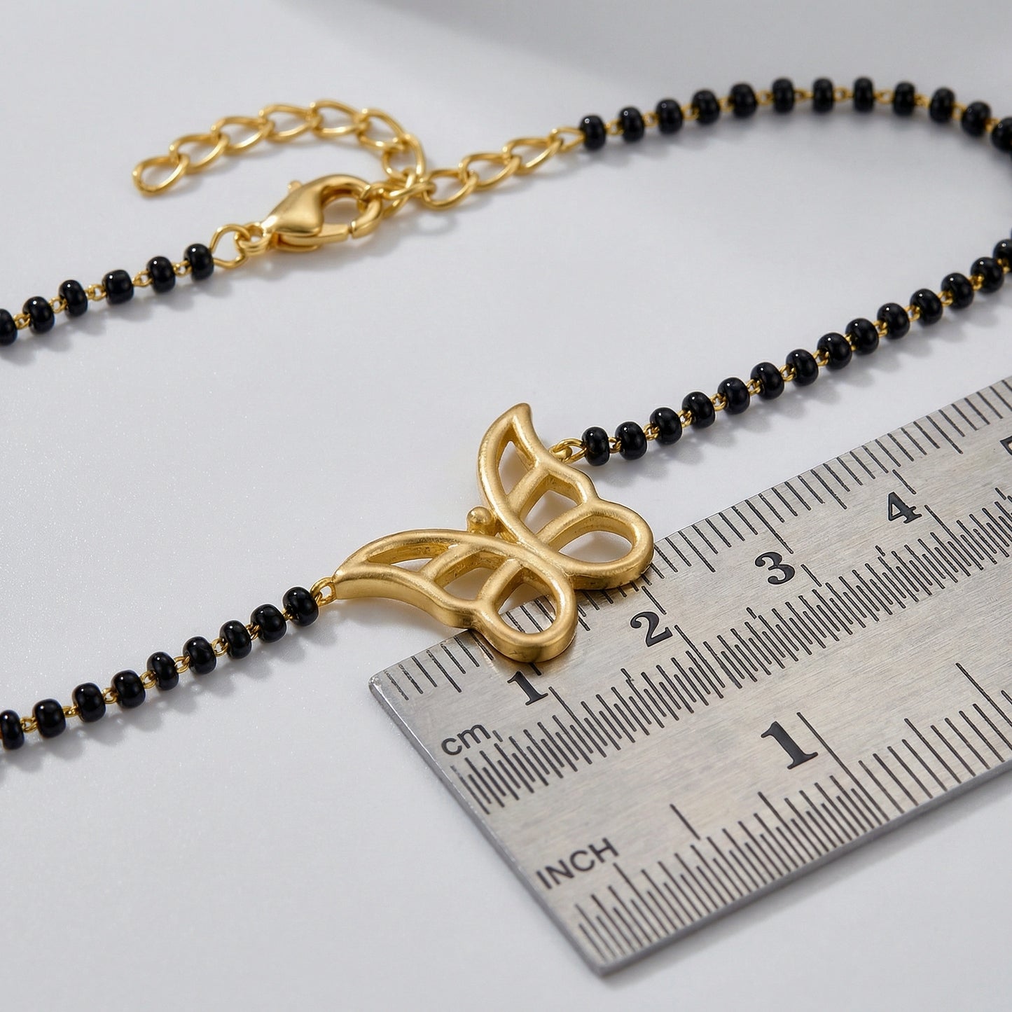 18k gold plated butterfly pendant on a black beaded chain with a ruler for scale on a light gray background