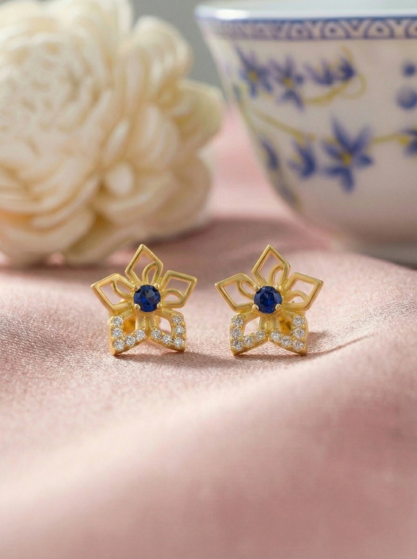18k gold plflower-shaped earrings with blue centers on a pink surface, with a blurred floral and teacup background.ted