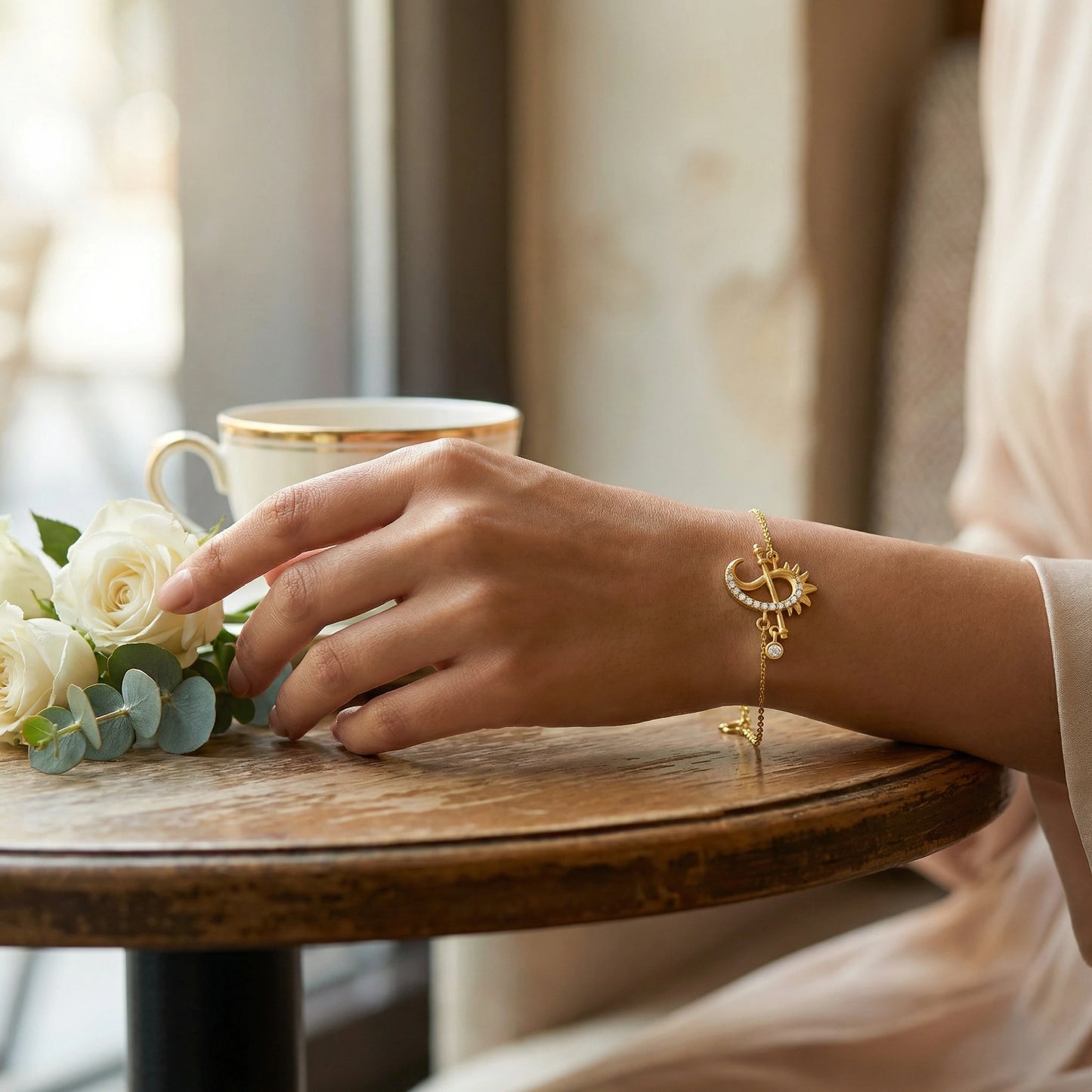 hand with 18k gold plated bracelet on a wooden table with flowers and a cup.