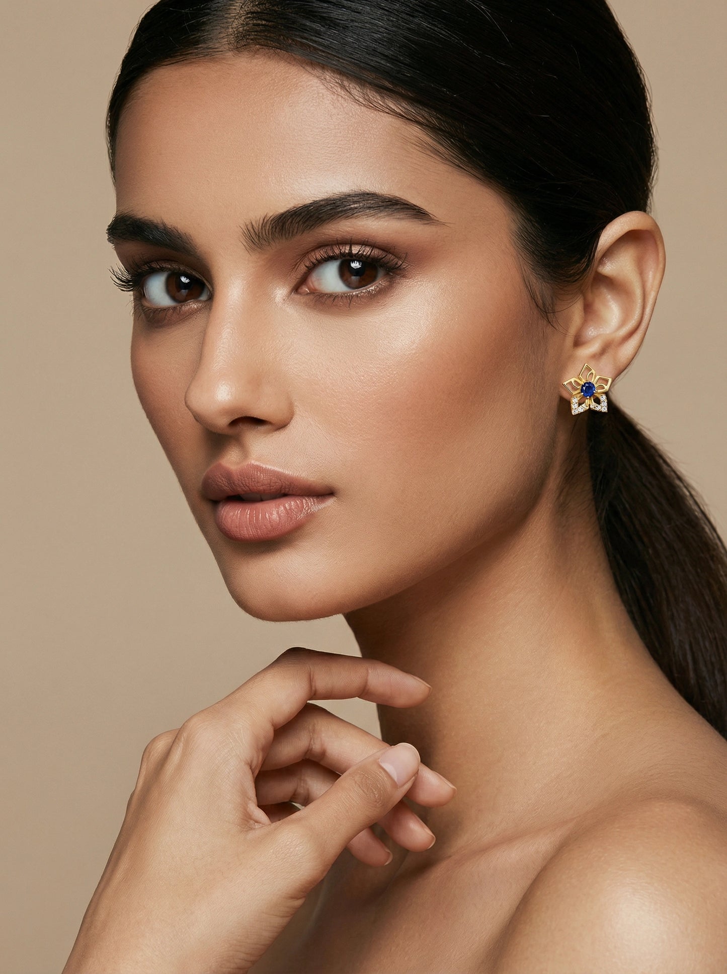 close-up of a woman wearing a 18k gold plated earring with a blue gemstone on a beige background
