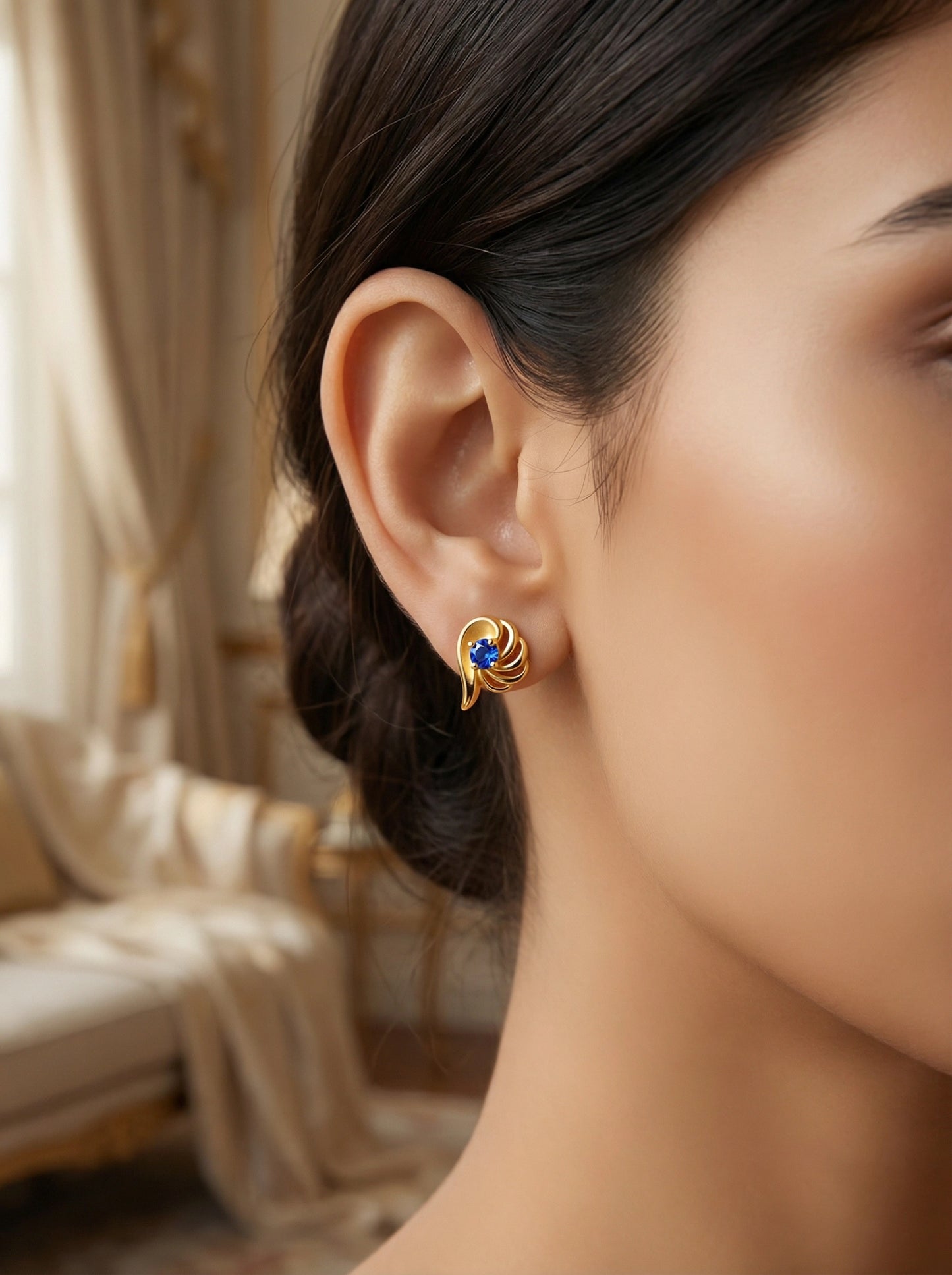 close-up of a woman wearing a 18k gold plated earring with a blue gemstone, blurred background
