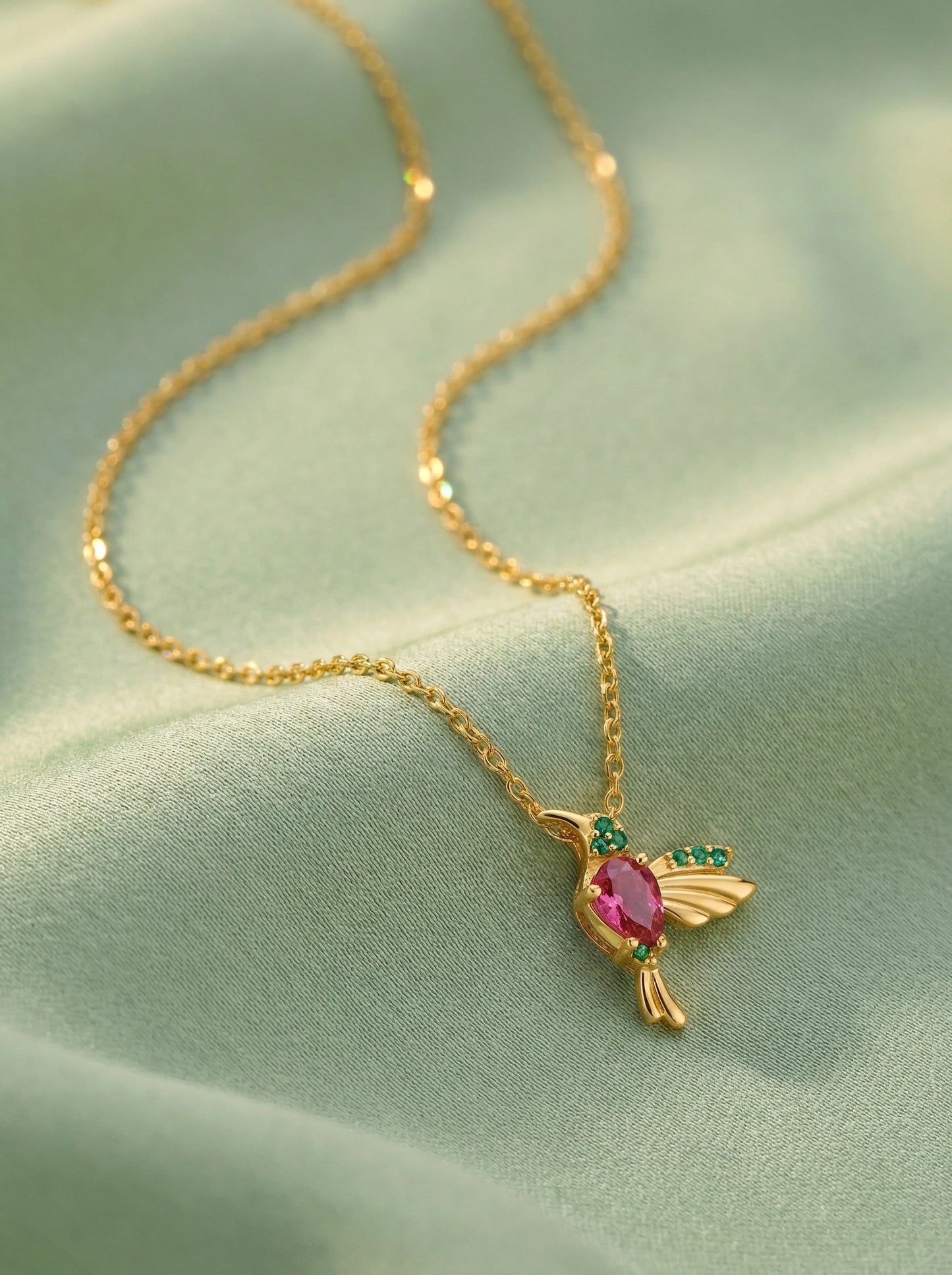 18k gold plated necklace with a bird-shaped pendant featuring a pink gemstone on a light green fabric background