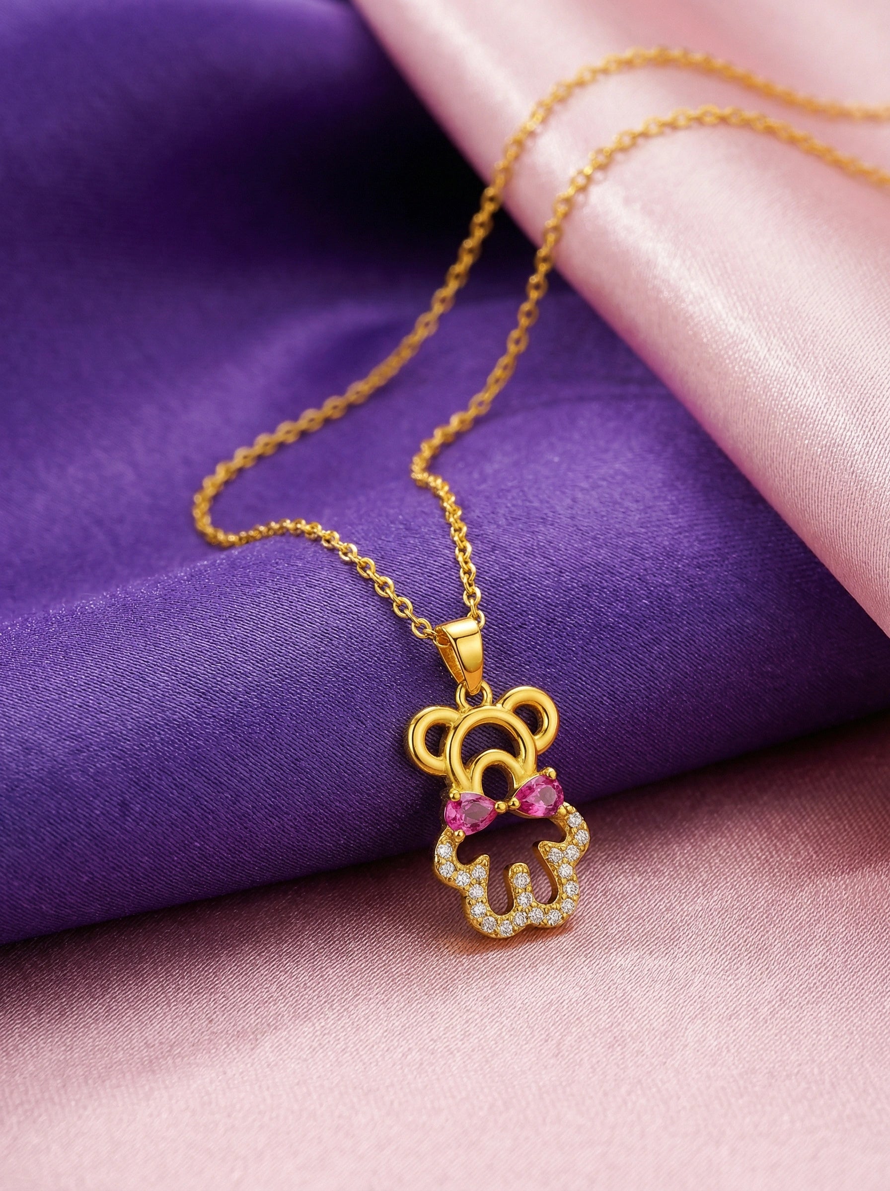 18k Gold plated necklace with a teddy bear pendant featuring pink and purple stones on a purple and pink fabric background.