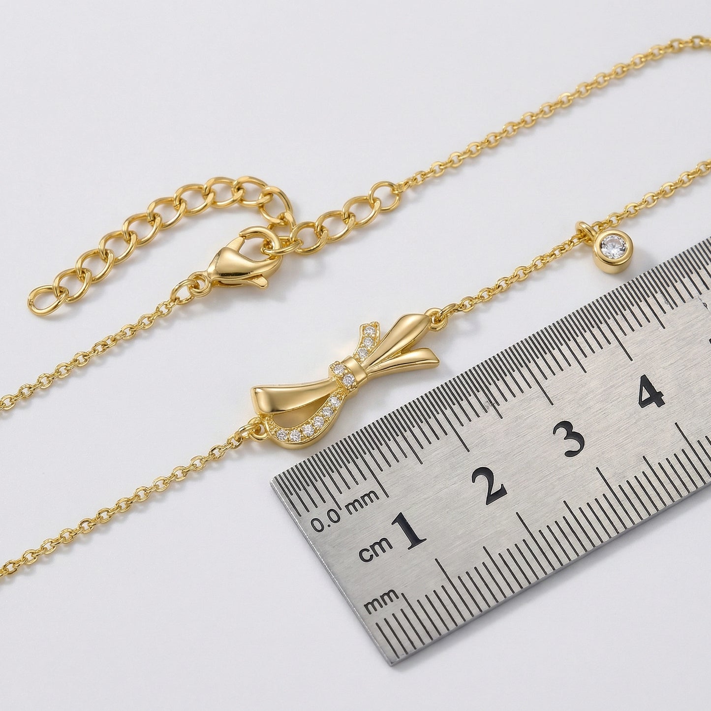18k gold plated bracelet with a bow charm next to a ruler for size reference on a white background